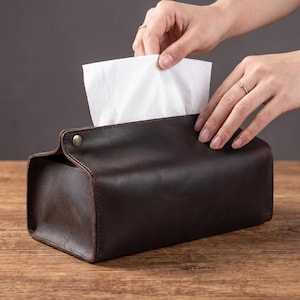 Może przedstawiać: Prostokątne etui na pudełko z chusteczkami wykonane z ciemnobrązowej skóry z widocznymi szwami na krawędziach i metalowym zapięciem na zatrzask z boku. Pojedyncza biała chusteczka papierowa jest wyciągana przez górny otwór.