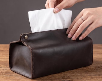 Könnte beinhalten: Ein rechteckiger Taschentuchbox-Bezug aus dunkelbraunem Leder mit sichtbaren Nähten an den Kanten und einem metallischen Druckknopfverschluss an der Seite. Ein einzelnes weißes Papiertaschentuch wird aus der oberen Öffnung gezogen.