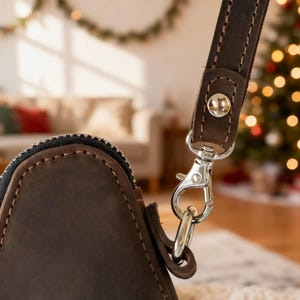 May include: Close-up of a brown leather bag with a silver-colored clasp and a matching strap. The bag has a zipper closure and is stitched with a lighter-colored thread. The background is blurred, with a Christmas tree and a couch visible.