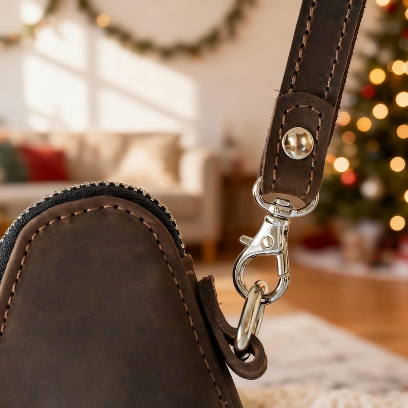May include: Close-up of a brown leather bag with a silver-colored clasp and a matching strap. The bag has a zipper closure and is stitched with a lighter-colored thread. The background is blurred, with a Christmas tree and a couch visible.