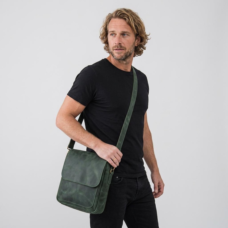 May include: A dark green leather messenger bag with a fold-over flap and an adjustable shoulder strap. The bag features a rectangular shape, a smooth matte finish, and metal hardware attachments for the strap, worn across the body.