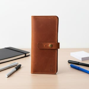 May include: A tall, rectangular, cognac-colored leather wallet featuring a vertical strap closure with a brass-toned metal snap button. The lower front of the wallet is embossed with the capital letters B and M stacked vertically.