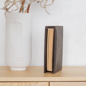 May include: A dark brown leather book cover standing upright on a light wood surface. The cover is wrapped around a book with visible cream-colored pages. The leather has a smooth, matte texture and clean, stitched edges along the spine and corners.