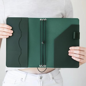 May include: Open green leather journal with multiple pockets and elastic closures. The journal is held open, showcasing the interior design. The cover is a darker shade of green, while the inside is a lighter green.
