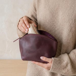 May include: A deep plum-colored leather pouch with a curved top zipper closure. The pouch has a smooth, matte finish and a structured base. A light-colored hairbrush with fine bristles is partially visible as it is being placed inside the pouch.