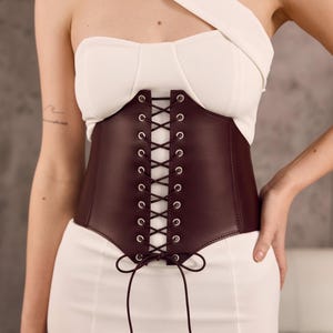 May include: A dark burgundy corset belt featuring a front lace-up design with silver-toned metal eyelets and matching thin cord laces tied in a bow at the bottom. The corset is constructed from a smooth material and worn over a white garment.