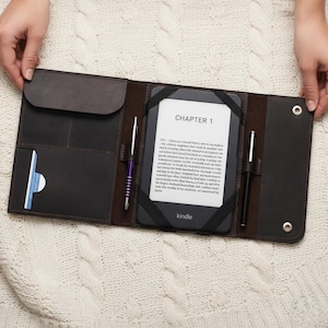 May include: A dark brown leather e-reader case featuring a central compartment for a Kindle device, secured by black elastic straps. The case includes a flap pocket, two pen loops with pens, and a card slot. The Kindle screen displays CHAPTER 1.