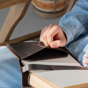 May include: A dark brown leather book cover featuring a smooth texture, a wrap-around strap closure, and an interior pocket flap. The cover is fitted around a thick book with cream-colored pages, showcasing visible stitching and natural leather grain.