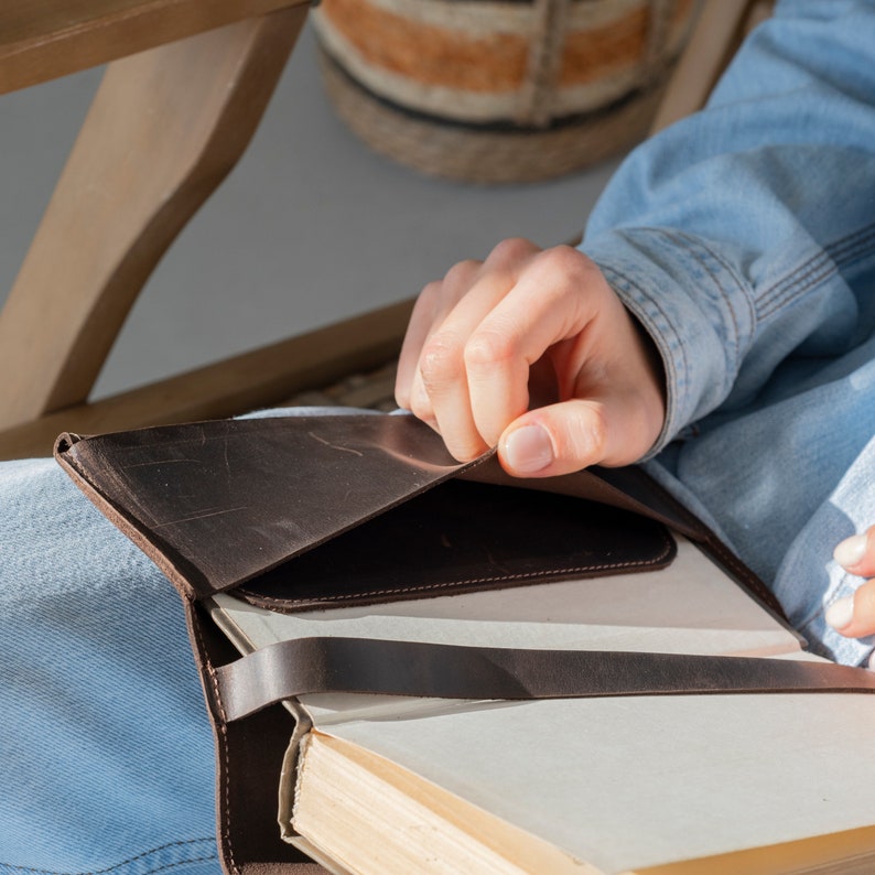May include: A dark brown leather book cover featuring a smooth texture, a wrap-around strap closure, and an interior pocket flap. The cover is fitted around a thick book with cream-colored pages, showcasing visible stitching and natural leather grain.