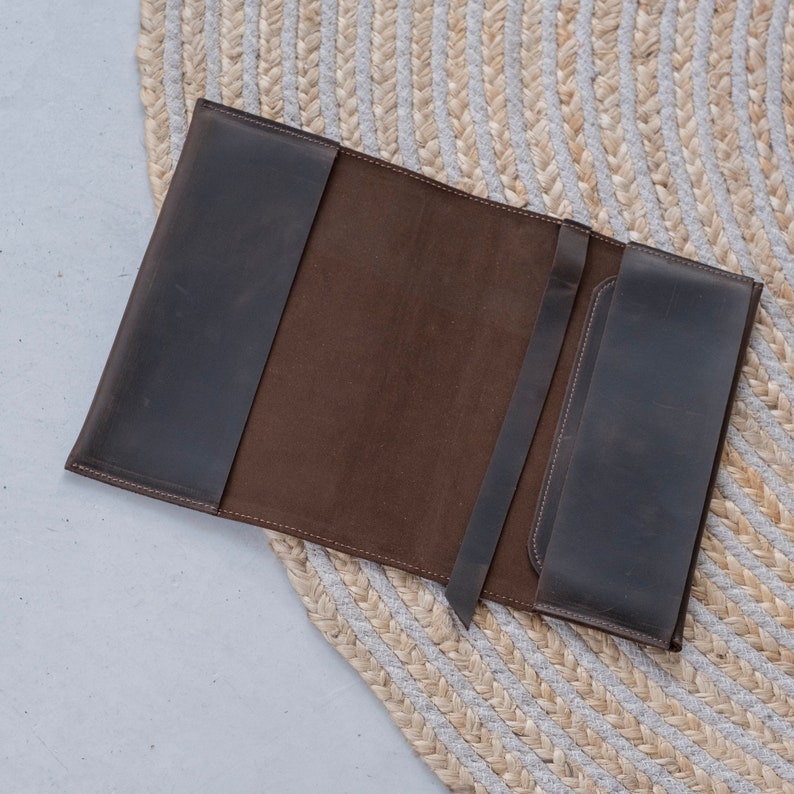May include: A dark brown leather book cover featuring two wide interior flaps for securing a book, a smooth central panel, and a thin vertical leather strap attached to the right side for closure, all constructed with visible tonal stitching.