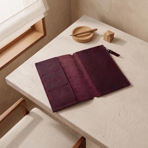 Puede incluir: Una carpeta de cuero de color morado oscuro con cierre de cremallera, que presenta un interior abierto con tres ranuras para tarjetas en el lado izquierdo y un bolsillo de longitud completa en el derecho, con textura mate y costuras visibles.