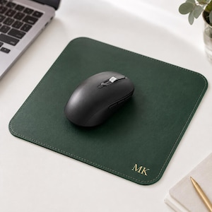 K&ouml;nnte beinhalten: Ein quadratisches, dunkelgr&uuml;nes Mauspad mit abgerundeten Ecken und goldenen, gepr&auml;gten Initialen MK in der unteren rechten Ecke. Eine schwarze, kabellose Computermaus mit Scrollrad und Seitentasten befindet sich in der Mitte des Mauspads.