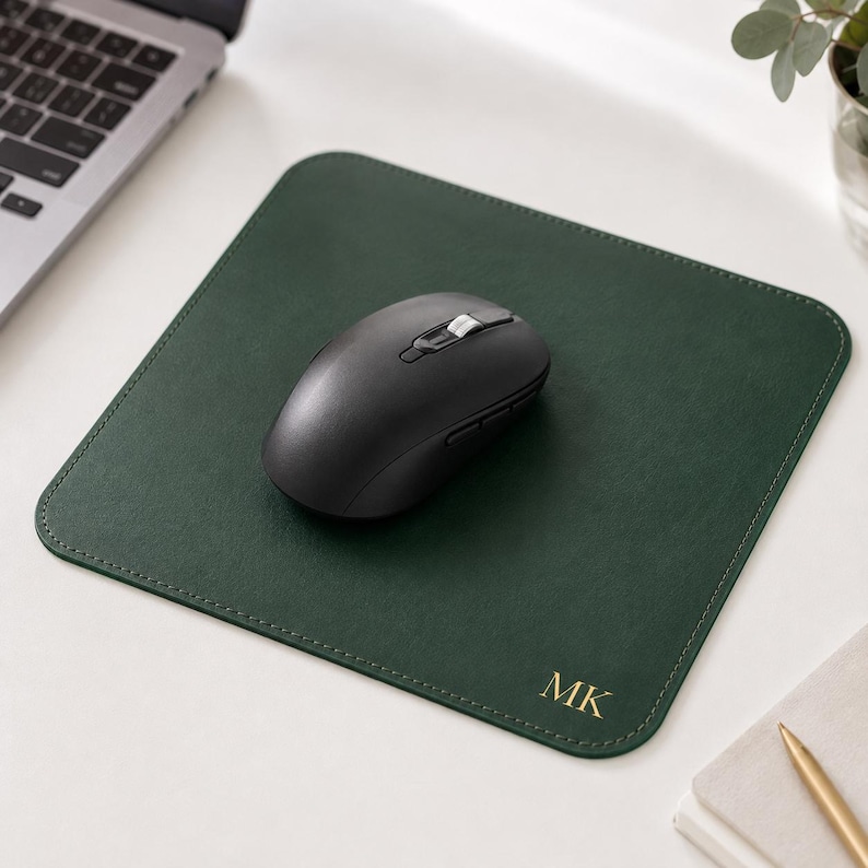 K&ouml;nnte beinhalten: Ein quadratisches, dunkelgr&uuml;nes Mauspad mit abgerundeten Ecken und goldenen, gepr&auml;gten Initialen MK in der unteren rechten Ecke. Eine schwarze, kabellose Computermaus mit Scrollrad und Seitentasten befindet sich in der Mitte des Mauspads.