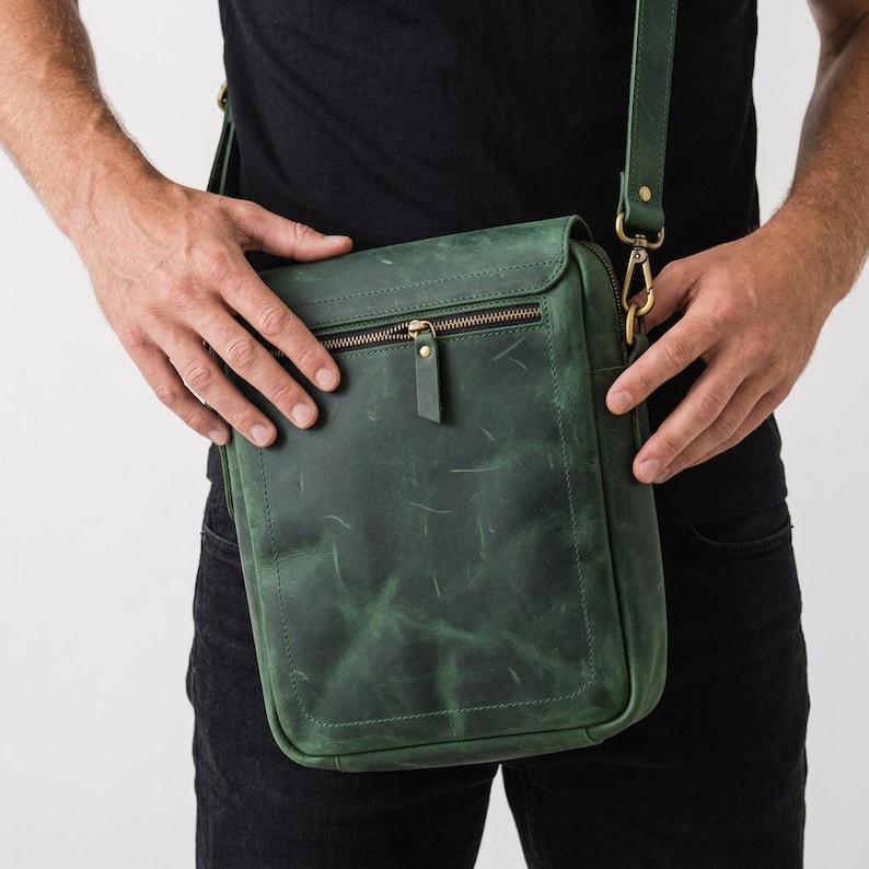 May include: A forest green leather messenger bag featuring a rectangular silhouette, a front zippered pocket with a matching leather pull tab, and antique brass hardware. The bag includes an adjustable shoulder strap attached with metal clips.