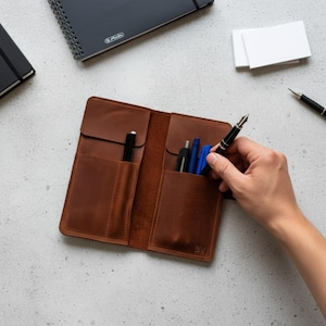 May include: A brown leather pen case with multiple vertical slots containing several pens. The case features a flap design and is embossed with the initials BV in the bottom right corner. The leather has a smooth, matte finish and visible stitching.