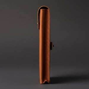 May include: A tan leather sleeve case shown from a side profile view. The case features a fold-over flap with a small metal stud closure and visible stitching along the edges, providing a slim and structured storage solution for flat items.