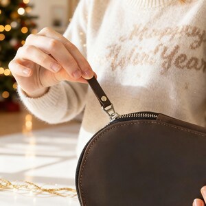 May include: A brown leather oval-shaped bag with a silver zipper and a small leather pull tab. The bag is being held by a person wearing a cream-colored sweater. The background includes a Christmas tree.