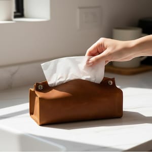May include: A brown leather tissue box with a curved top and silver rivets. White tissues are being pulled from the opening. The box is rectangular and sits on a white countertop. The background is a kitchen setting.