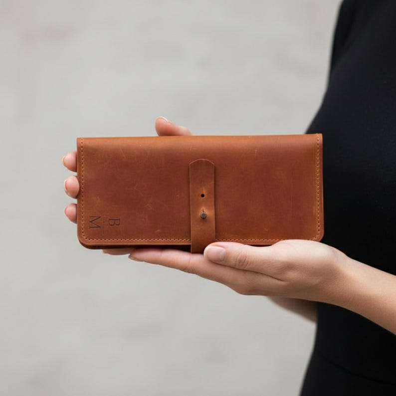 May include: A rectangular tan leather wallet featuring a smooth finish, a central vertical strap closure with a single metal stud, and the embossed letters B and M on the bottom left corner. The wallet has visible stitching along the edges.