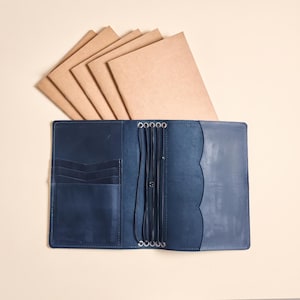 May include: A navy blue leather traveler's notebook cover featuring internal card slots, a large pocket, and multiple elastic bands for securing inserts. Five tan paper notebooks are fanned out behind the open leather cover.