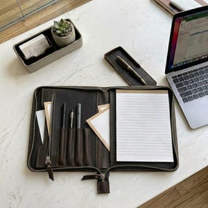 May include: A dark brown leather zippered portfolio organizer featuring an interior lined with pockets for pens, a zippered compartment, and slots for documents. A white lined notepad is secured on the right side, and the case is open to display its contents.