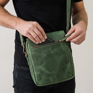 May include: A forest green leather crossbody bag featuring a rectangular shape, a top flap with a zippered pocket, and antique brass hardware. The bag includes a matching adjustable shoulder strap attached with metal clips and visible stitching.