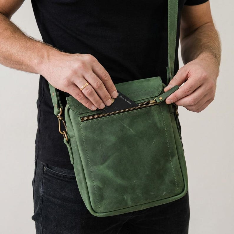 May include: A forest green leather crossbody bag featuring a rectangular shape, a top flap with a zippered pocket, and antique brass hardware. The bag includes a matching adjustable shoulder strap attached with metal clips and visible stitching.