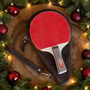 May include: A red ping pong paddle with a white handle and a brown case. The paddle is inside a brown leather case with a wrist strap. The image is decorated with Christmas ornaments and greenery.