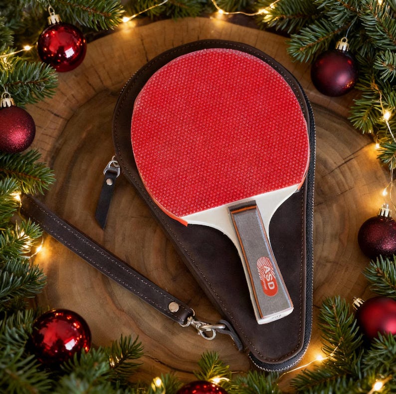 May include: A red ping pong paddle with a white handle and a brown case. The paddle is inside a brown leather case with a wrist strap. The image is decorated with Christmas ornaments and greenery.