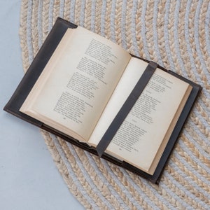 May include: An open hardcover book with aged, cream-colored pages featuring printed Russian text. The book is encased in a dark brown leather cover with a matching leather strap that secures the pages in place across the center of the open spread.