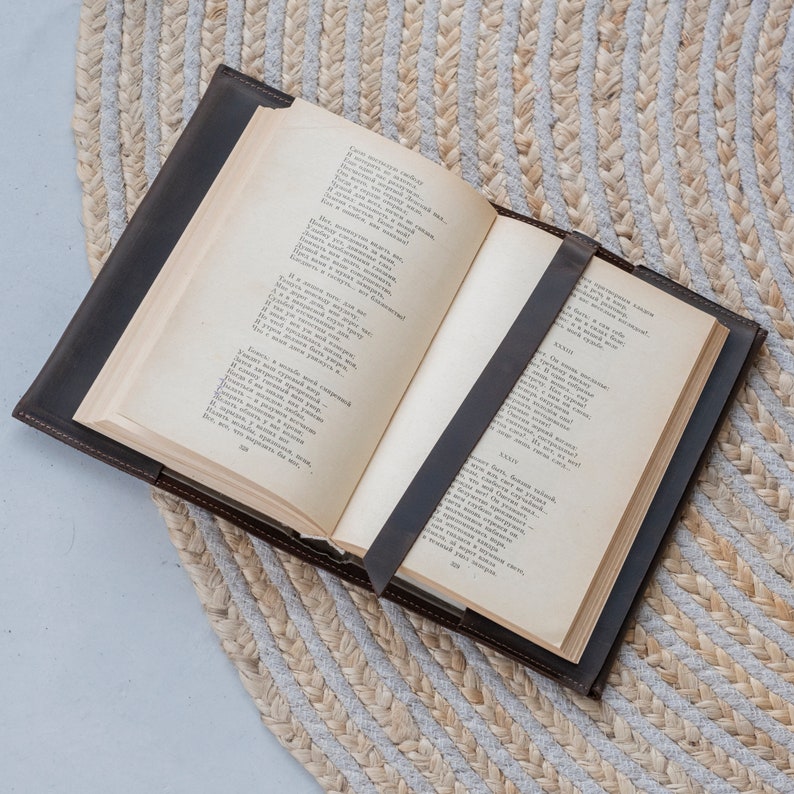 May include: An open hardcover book with aged, cream-colored pages featuring printed Russian text. The book is encased in a dark brown leather cover with a matching leather strap that secures the pages in place across the center of the open spread.