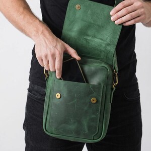 May include: A forest green leather crossbody bag featuring a rectangular silhouette, a fold-over flap with a magnetic snap closure, and an external front pocket. The bag includes brass-toned metal hardware and an adjustable shoulder strap.