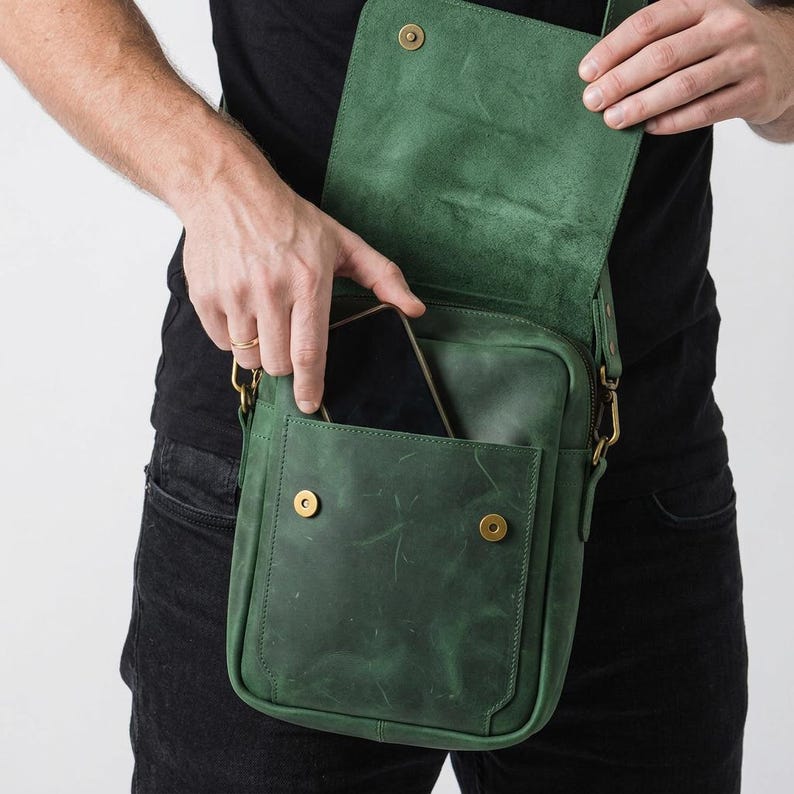 May include: A forest green leather crossbody bag featuring a rectangular silhouette, a fold-over flap with a magnetic snap closure, and an external front pocket. The bag includes brass-toned metal hardware and an adjustable shoulder strap.