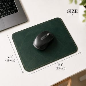 K&ouml;nnte beinhalten: Ein rechteckiges Mauspad aus dunkelgr&uuml;nem Leder mit sichtbaren N&auml;hten an den R&auml;ndern, auf dem eine schwarze kabellose Computermaus liegt. Die Ma&szlig;e sind deutlich mit 18 cm mal 23 cm angegeben und bieten eine klare Skalierung f&uuml;r das Produkt.