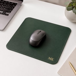 K&ouml;nnte beinhalten: Ein quadratisches, dunkelgr&uuml;nes Mauspad mit abgerundeten Ecken und goldfarbenen Monogrammbuchstaben MK in der unteren rechten Ecke. Eine dunkelgraue, kabellose Computermaus befindet sich in der Mitte der glatten, matt texturierten Oberfl&auml;che.