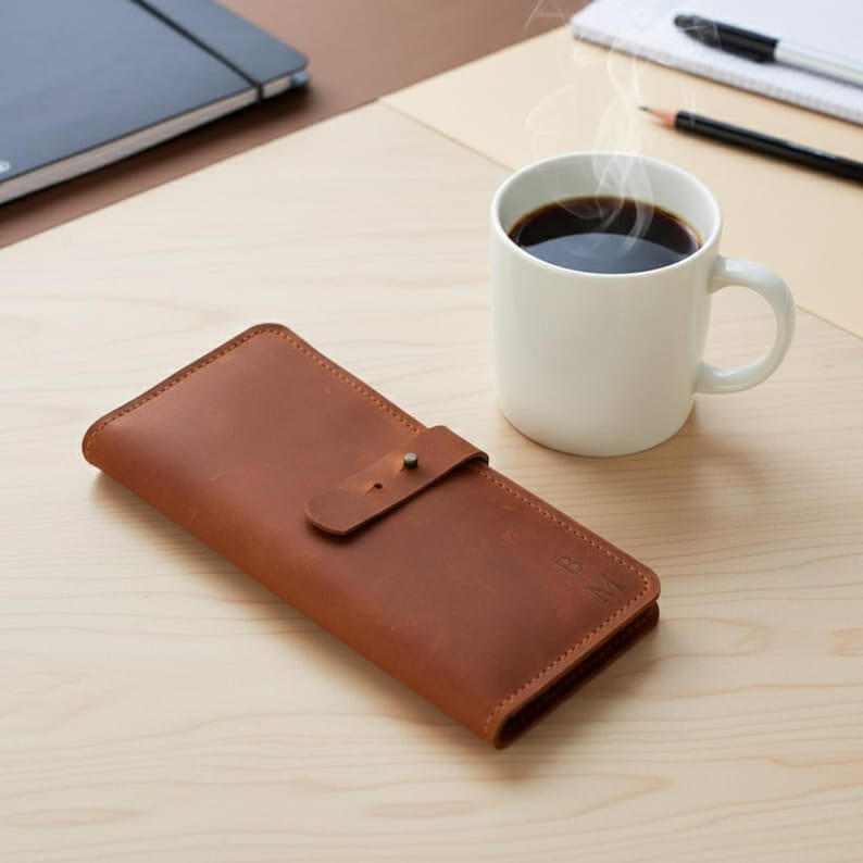 May include: A rectangular tan leather wallet featuring a fold-over flap with a single metal stud closure. The wallet has a smooth, matte finish and includes the embossed initials B M on the bottom right corner of the front exterior surface.