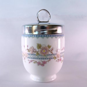 Peut inclure: Un coquetier en céramique blanche avec un motif floral et un couvercle argenté avec une poignée en forme d'anneau.