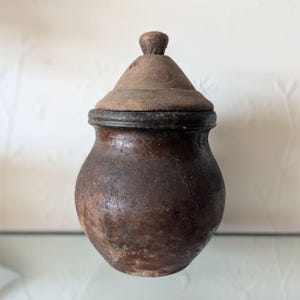 May include: A small, round, dark brown earthenware pot with a textured, weathered surface and a wide opening. The container features a removable, conical wooden lid with a small, rounded knob handle at the very top, creating a rustic appearance.