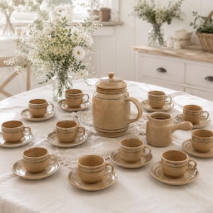 Op de afbeelding: Een beige keramische theeset met een theepot, melkkan en meerdere kopjes en schotels, gerangschikt op een ronde tafel. De set heeft een rustieke, vintage uitstraling, met een eenvoudig ontwerp en een lichtbruine kleur. De tafel is bedekt met een wit tafelkleed.