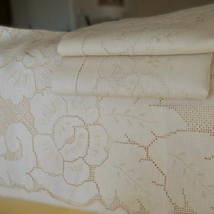 May include: A set of three white linen tablecloths with delicate embroidered floral designs. The tablecloths are layered on top of each other, creating a luxurious and elegant look.