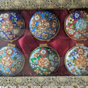 May include: A set of six round metal trinket boxes with hinged lids, each featuring intricate floral cloisonné enamel patterns in vibrant colors including blue, red, green, and white, displayed inside a rectangular box with a patterned frame.