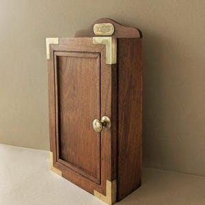 Könnte beinhalten: Ein hölzerner Schlüsselkasten mit Messing-Eckakzenten und einem kleinen Messingknauf. Das Wort "KEYS" ist auf einer kleinen Messingplatte über der Tür eingraviert. Der Schrank ist rechteckig und hat eine dunkelbraune Oberfläche.