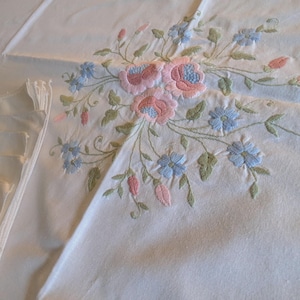 Pode incluir: Toalha de mesa bordada creme com um desenho floral. O bordado inclui flores rosa e azuis com folhas verdes. A toalha de mesa tem uma borda plissada no lado esquerdo.
