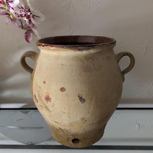 May include: A large, round, beige ceramic vessel with two side handles and a dark brown interior rim. The exterior surface features a weathered, textured finish with scattered rust-colored spots and a small circular hole near the base.
