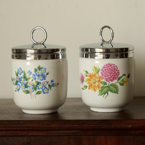 May include: Two white ceramic egg cups with silver lids and floral designs. One cup features blue forget-me-not flowers and the other has yellow and pink flowers.