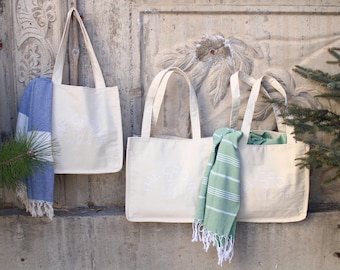 May include: Four white canvas tote bags with fabric straps hanging on a wall. Two of the bags have blue and green striped towels hanging from them.