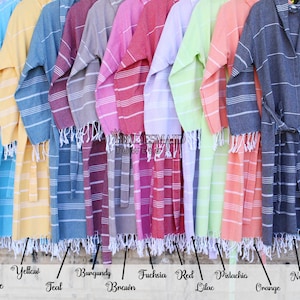 May include: A row of ten Turkish towels, each with a different color stripe pattern. The colors are: royal turquoise, yellow, teal, burgundy, fuchsia, red, pistachio, orange, and navy. The towels are all hanging on a metal fence.