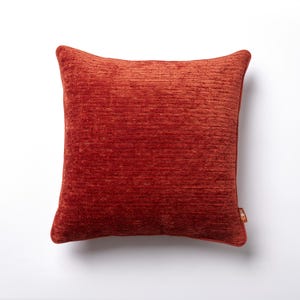 May include: A square, rust-colored decorative pillow with a textured surface. The pillow has a soft, plush appearance and a tag on the lower right corner. The pillow is likely used for home decor.