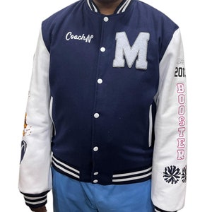 Può includere: Una giacca da baseball blu navy e bianca con una grande "M" bianca sul davanti. Le maniche sono bianche con un "BOOSTER" rosa e un design a pompon di cheerleader. La giacca ha una striscia bianca intorno al fondo e ai polsini. Il testo "Coach N" è ricamato sul lato sinistro della giacca.