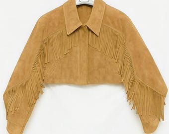 May include: A tan suede cropped jacket with fringe detailing along the sleeves and chest. The jacket features a collared neckline and button closures. The sleeves are long and end in a rounded cuff. The jacket is hanging on a white hanger.